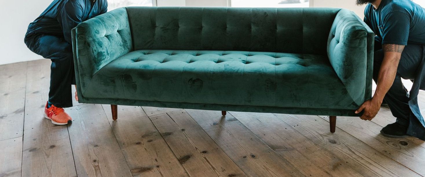 Two men carrying a green sofa in a sunlit room with wooden floors and arched windows.